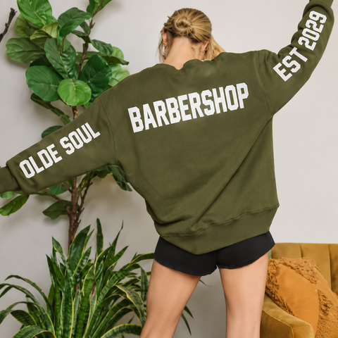 A woman wearing an oversized olive green sweatshirt with 'OLDE SOUL BARBERSHOP EST 2029' printed in white across the back, paired with black shorts and black fringe boots, posed in a living room setting with indoor plants and a mustard yellow couch.
